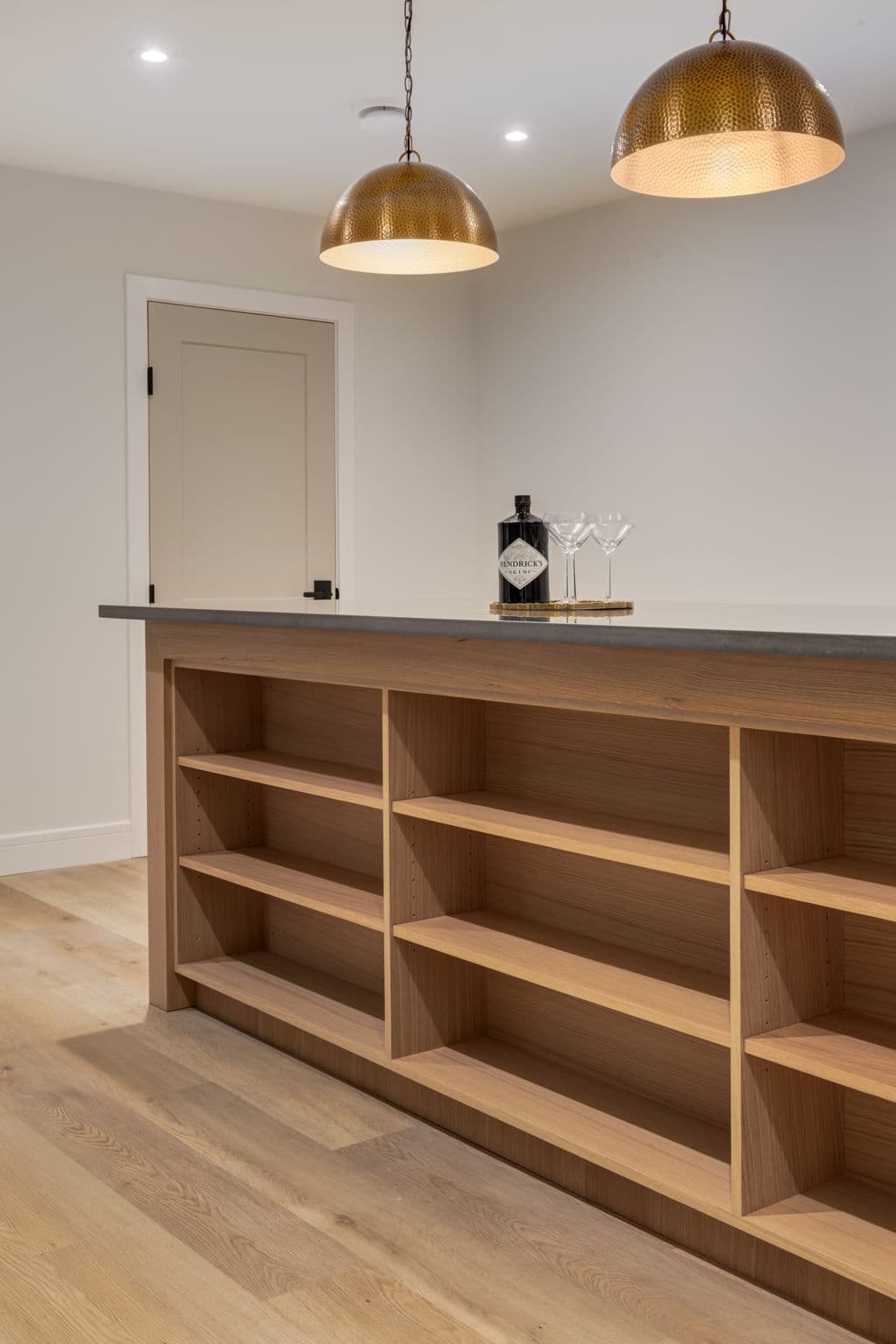 Lower-level bar with open white oak shelving and brass pendant lights in the Varal Road renovation by Platinum Build Homes in Varsity, Calgary.